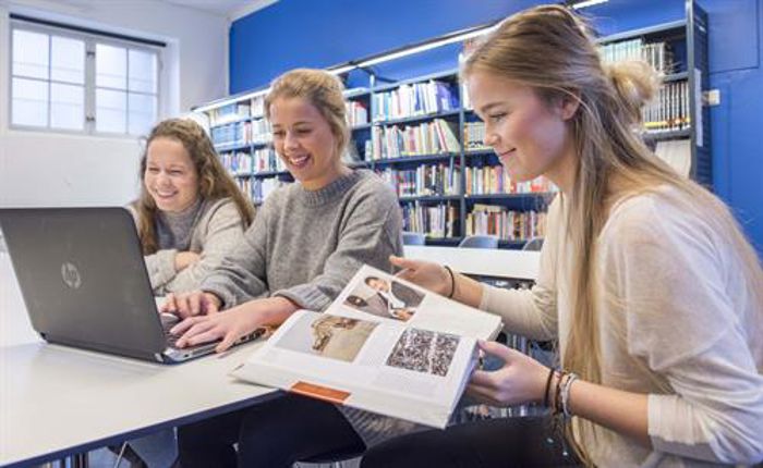 3 jenter ved et bord, som ser på en pc og leser i en lærebok. Bokhyller og et vindu i bakgrunnen